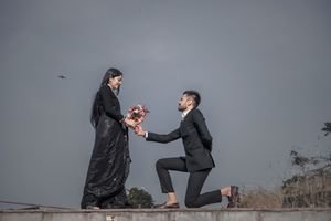 Man proposing with a bouquet, kneeling before a woman in a black dress outdoors.